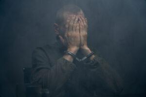 man-sitting-on-chair-with-handcuffs / arrested-suspect-covering-face-in-dark-foggy-room