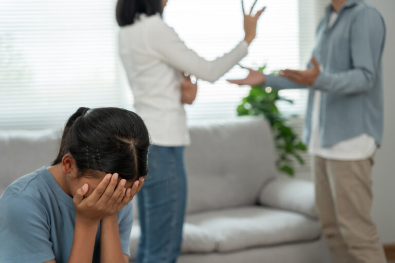 Young girl covering her face with her hands and parents with H1B Visa applications quarreling in the background.