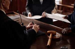 A judge signing legal documents in a courtroom while two attorneys present paperwork.